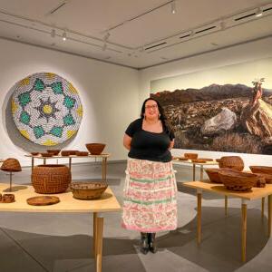 Mer和a Roberts st和s in the middle of a ring of Cahuilla baskets sitting on curved tables. In the background are installations of a basket made of cans 和 a scenic photograph of a desertscape.